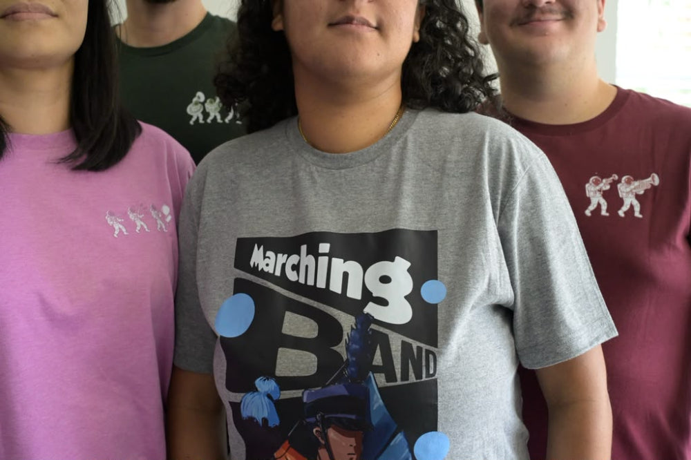 Three people standing together wearing graphic t-shirts. The central person wears a heather gray t-shirt with a black graphic that reads 'Marching Band' and blue circular accents.